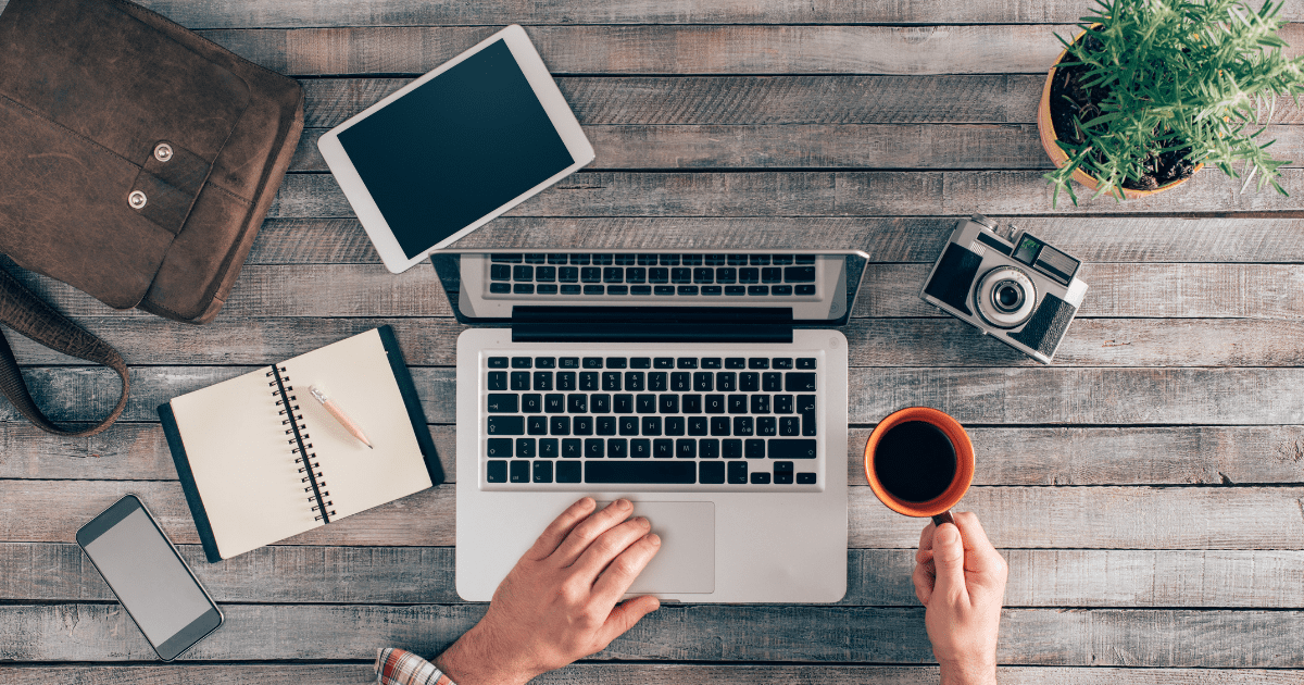 5 tips for working, planning or studying on the move Workspace with hands on laptop and coffee, on a wooden table with plant, camera, smartphone, open notebook, tablet and bag, to show working or studying remotely or for a distributed team and promote concepts like mind mapping and online chat for team connectivity